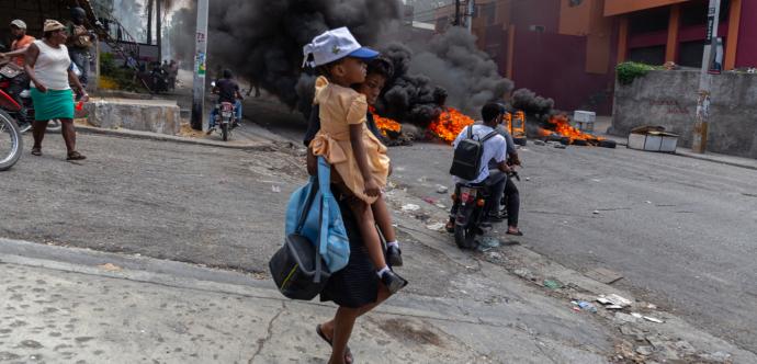 Une mère tient son enfant et des sacs, fuyant un arrière plan de feu et de fumée