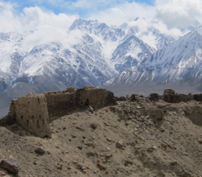 L'image montre un paysage accidenté et époustouflant d'un village pamiri niché dans les montagnes du Pamir, au Tadjikistan. Les ruines d'anciennes structures en pierre sont visibles au premier plan, perchées sur un terrain sec et rocailleux. À l'arrière-plan, les imposants pics enneigés de la chaîne du Pamir s'élèvent de façon spectaculaire dans un ciel bleu partiellement nuageux.