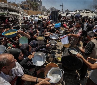 Des bénévoles distribuent de la nourriture à une foule dense de personnes, femmes, hommes et enfants, tendant des casseroles, bols et seaux dans une zone urbaine détruite. L’image reflète une situation d’urgence humanitaire, avec des bâtiments en ruine et des signes de conflit à l’arrière-plan.