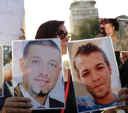 Un groupe de personnes manifeste en tenant des photos de proches disparus. Trois portraits d’hommes souriants ou sérieux sont clairement visibles, protégés par du plastique. À l’arrière-plan, d’autres participants tiennent des pancartes avec du texte en arabe. La scène se déroule en extérieur, en plein jour.