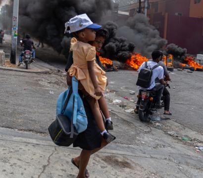 Une mère tient son enfant et des sacs, fuyant un arrière plan de feu et de fumée
